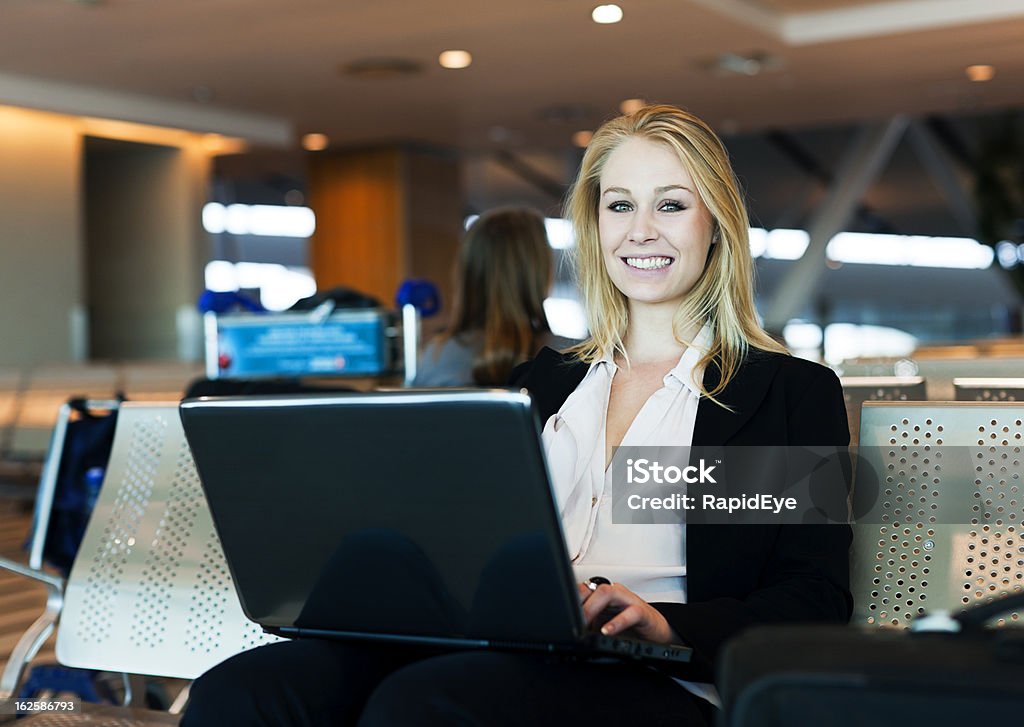 Blonde Femme d'affaires à l'aide d'ordinateur portable à l'aéroport, souriant looks concourse - Photo de Adulte libre de droits