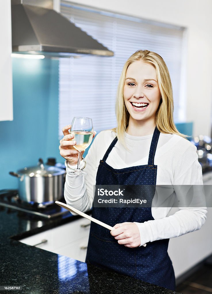 Bionda sorridente cottura in cucina con il vino bianco e bevande - Foto stock royalty-free di Cucinare