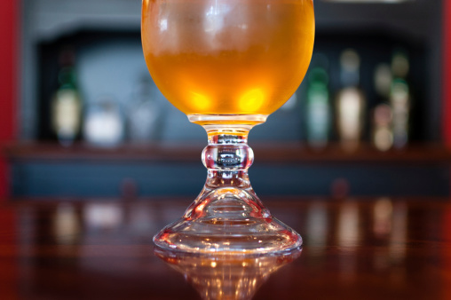 A goblet of golden beer is shot against a red background. The glass is positioned on a glossy cherry wood table. In the background, wine glasses and wine bottles can be seen. Wonderful reflections fall onto the table, from both the background elements and the goblet of beer.