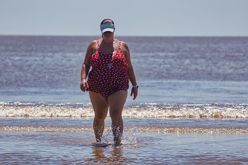 Full figured woman in swimsuit walking along beach splashing in ocean water waves or tide pool during summer vacation or road trip.