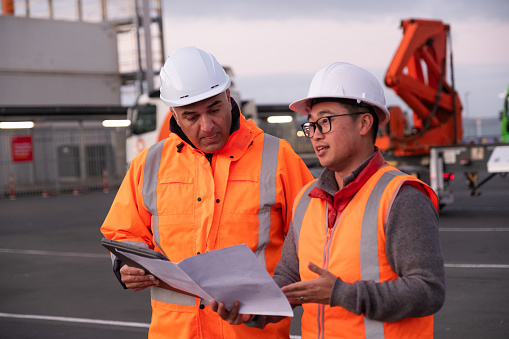 Two males wearing construction clothing having a discussion while looking at paper documents