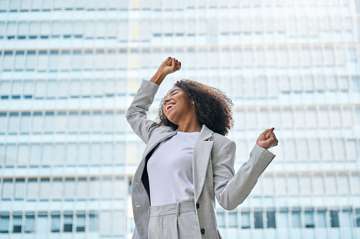 Happy excited confident professional young African American business woman office leader executive wearing suit celebrating financial goals standing in big city street feeling success and freedom.