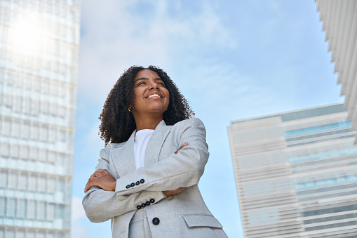 Happy smiling confident professional young African American business woman office leader executive wearing suit standing in big city on street arms crossed thinking of success, feeling proud outdoors.