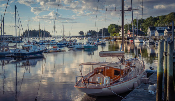 A mix of fishing and leisure boats at anchor in Camden Harbor. The sun rises to the east. The water is calm with cool clouds reflecting in the sky.