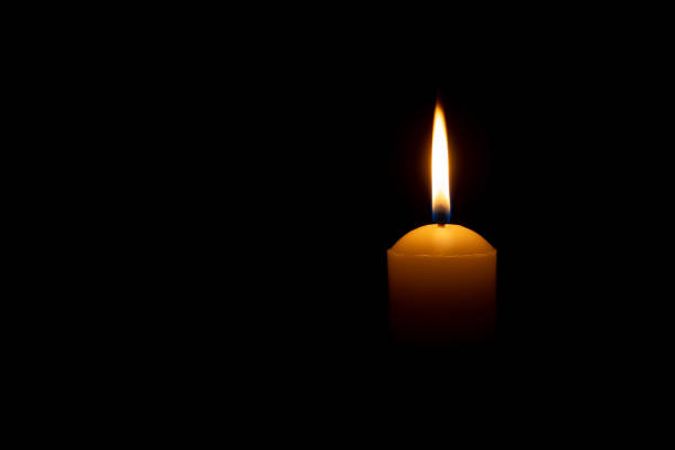 istock Single burning candle flame or light glowing on a big yellow candle on black or dark background on table in church for Christmas, funeral or memorial service with copy space. 1626397867