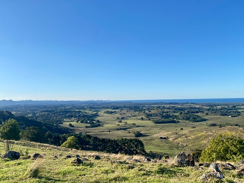 Horizontal landscape from high up of grassy valley side hill looking out to ocean horizon with trees and crops under a clear blue sky in Byron Bay Australia