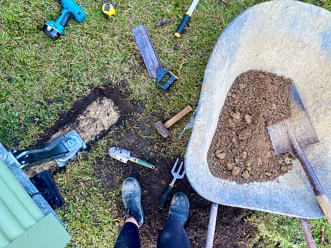 Horizontal flat lay grass yard with gravel dirt in wheel barrow shovel and garden and building tools and leg and boot on grass area as part of building frame for Tiny House Land Preparation  in country Australia