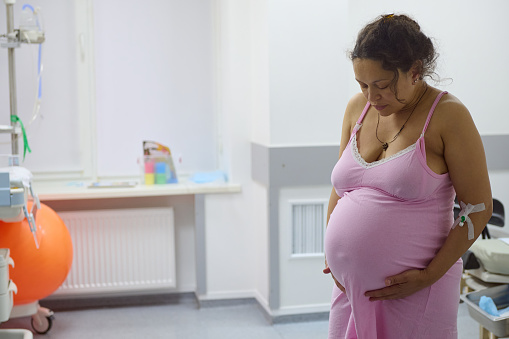 Middle aged multi-ethnic pregnant woman, birthing mother with painful contraction, at hospital ward, touching belly and looking down. Childbirth process at maternity hospital. Pregnancy 40 week