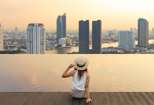 Pretty tourist woman is relaxing and enjoying her rooftop swimming pool view with cityscape background for vacation and travel at the sunset with skyscraper and downtown highrise condominium