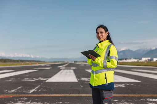 Happy Asian female airport worker looking at the camera and posing with her digital tablet on the airport runway.