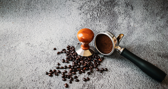 Equipment in a coffee shop of barista coffee tool portafilter with tamper and dark roasted coffee beans on gray background