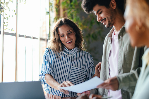 A smiling Caucasian female holding a piece of paper and showing something on it while talking to her coworker.