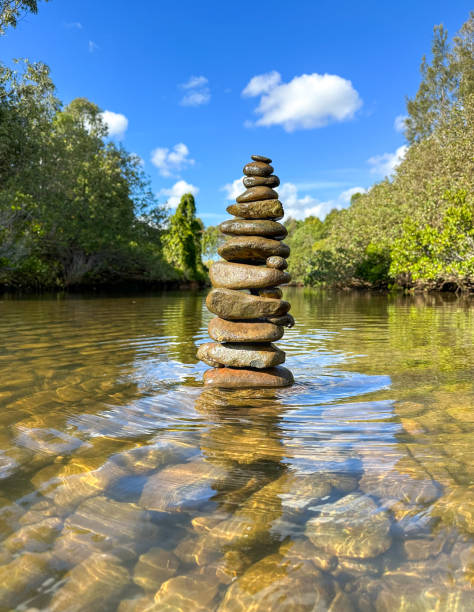 Rock stack Cairn built in the Nerang River.  Can see rocks under water.  Trees either side.  Blue skies in the distance.  Gold Coast Queensland Australia