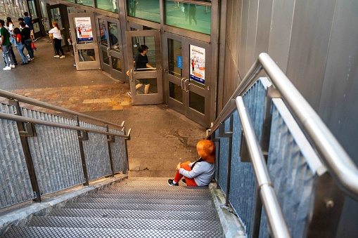 Woman in orange pants and a hat sits at the bottom of metal staircase steps at the Staten Island Ferry in New York City, public entrance at bottom with commuters going in and out the doors. With copy space.