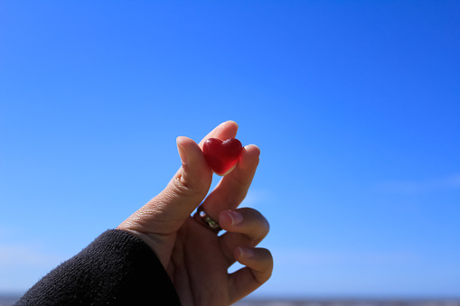 A woman hand holding a heart shape red gummy with the blue sky and beach scene in the background. In Crosby Beach in Liverpool, UK, in Merseyside county of North West England. For relationship concept