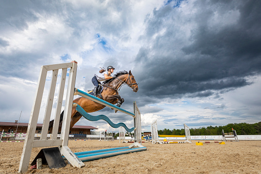 Illuminated by sunlight a female show jumper on her brown mare is crossing over the hurdle in front of a thunderstorm when jumping during a horse riding work shop at Ranshofen Horse Riding Arena, Austria.\nCanon EOS 5D Mark IV, 1/1000, f/5,6 , 24 mm.