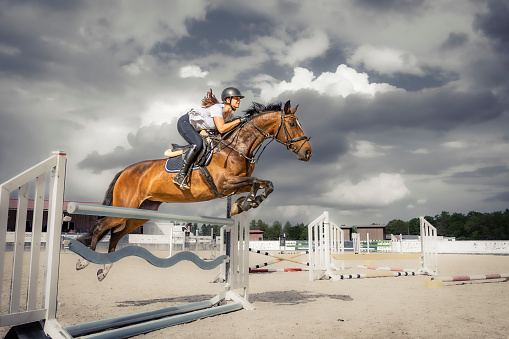 Illuminated by sunlight in front of a thunderstorm a female show jumper on her brown mare is crossing over the hurdle when jumping during a horse riding work shop at Ranshofen Horse Riding Arena, Austria.\nCanon EOS 5D Mark IV, 1/1000, f/5,6 , 24 mm.