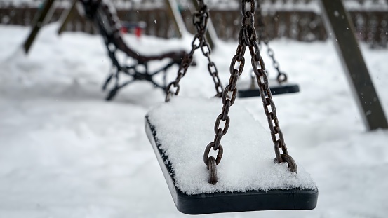 Closeup of snow slowly covering empty chain swings on winter children playground at park.