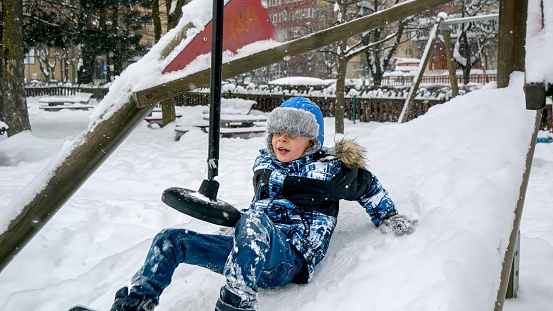 Little boy falling on the playground covered with snow from the zipline. Children playing and having fun outdoors during winter holidays and weekend.