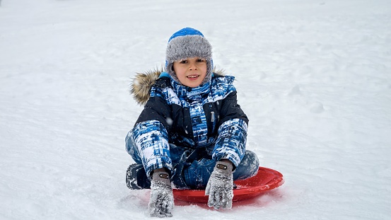 Portrait of happy cheerful boy sitting on plastic sleds and smiling in camera during snowfall.