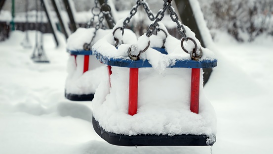 Closeup of empty chain swings on public playground at park being covered with snow during snowfall. Concept of winter holidays, bad weather and blizzard.