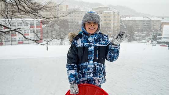 Happy smiling boy holding his plastic sleds on a snowy hill at snowfall. The concept of children having fun during winter, Christmas holidays, and playing outdoors in the snow