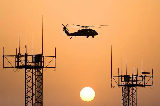 UH-60 Blackhawk helicopter flying between two radio towers, silhouetted against and orange sky and the setting sun.