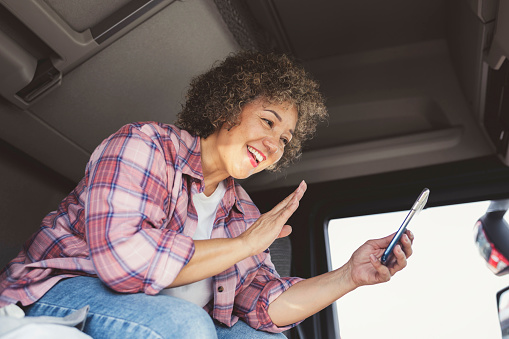 Smiling  Mixed Race Female Truck Driver Using Her Phone and Waving in the Lorry Cabin During a Break