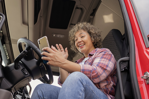 Smiling Mixed Race Female Truck Driver Using Her Smartphone in the Lorry Cabin During a Break