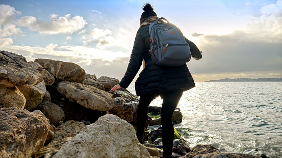 Young adventurous woman hiking and exploring the rugged cliffs along the sea coast during a cold winter sunset. A concept of tourism, travel, and winter vacation