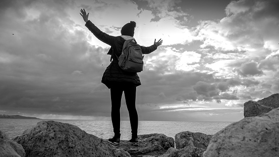 Black and white photo of a female hiker stands on the cliff edge at the sea coast, arms stretched out enjoying view of cold ocean waves. Perfect for adventure, nature relaxation, exploration, and travel