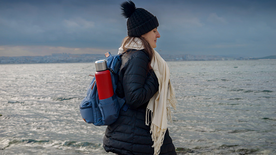 Female hiker, equipped with a backpack and thermos, exploring a rocky sea beach on a cold, cloudy day. Addition to any travel, tourism or adventure-themed project