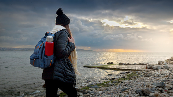 Young smiling woman with backpack and thermos walking along the rocky sea beach, enjoying the cool breeze and the sound of the waves. Concept of adventure, exploration, and travel