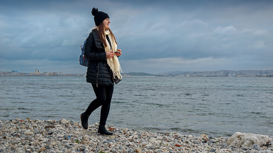 Smiling woman in hat and scarf walking on ocean beach and collecting beautiful sea shells. Concept of hiking, travel, exploration, tourism and journey
