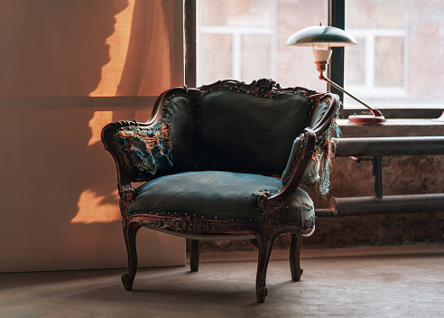 Classic vintage armchair with beautiful carved wooden elements and blue torn upholstery in grunge room with green lamp on windowsill. Retro interior concept with copy space.
