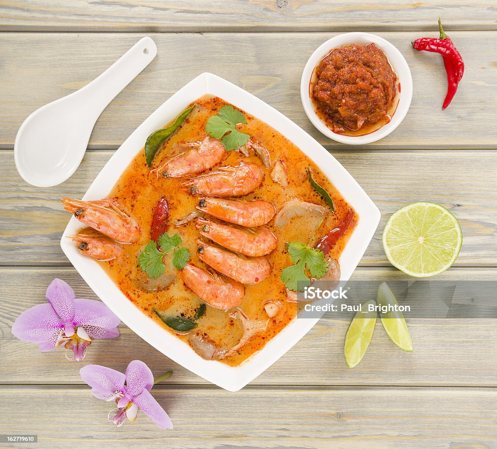 Overhead view of bowl of Tom Yum Nam Khon on a wooden table Creamy Thai soup with prawns and mushrooms garnished with coriander leaves and served with lime wedges. Coconut Milk Stock Photo