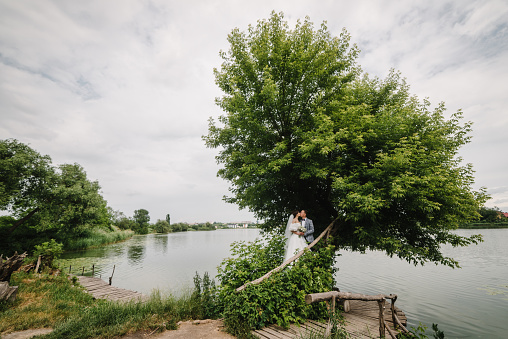 The bride and groom kissing and hold hands in nature. Newlyweds couple are standing near a big tree near lake. A love story country. Happy wedding day of marriage. Getting married outdoor.