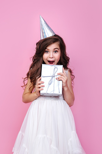 Portrait of surprised kid girl n birthday party princess hat holding gift box on a pink background