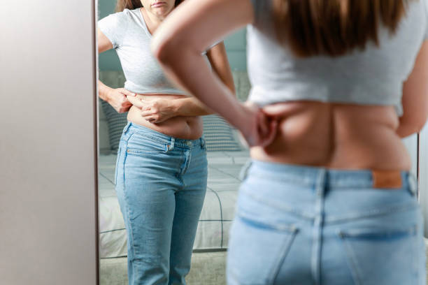 istock Young woman touching her belly looking in mirror at home 1627327596