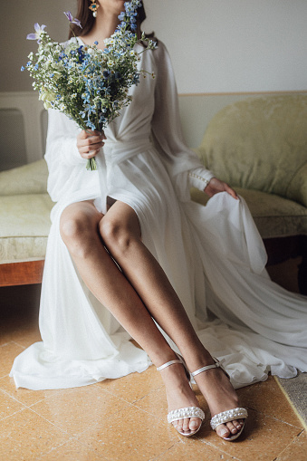 Young brunette wearing elegant white dress with veil and golden shoes standing with bouquet