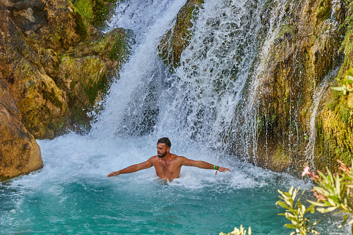 Les Fonts de l'Algar, Alicante, Spain - August 13, 2023: A young man cools off in the waterfall of a cold mountain river on a hot summer day.