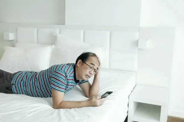 A lone man lying in bed, lost in thought as he stares at his smartphone.