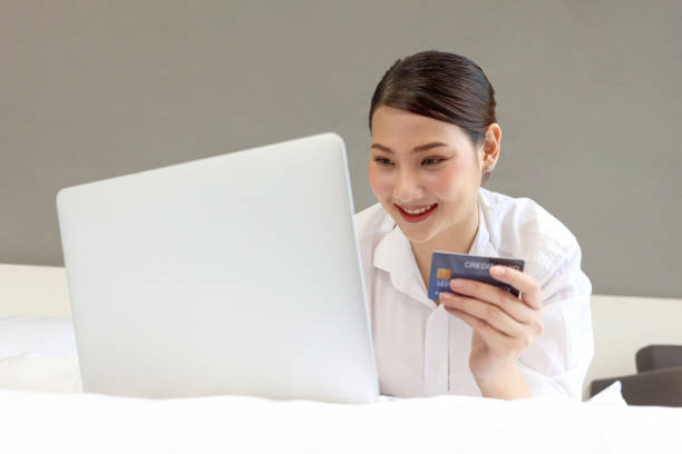 istock Young happy beautiful Asian woman using laptop computer and holding credit card while lying on the bed, businesswoman working online at home, e-commerce, internet banking, online shopping easy concept 1627409084