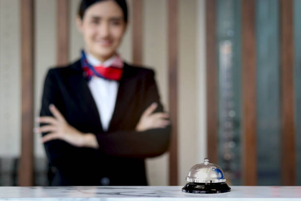 istock Silver bell on hotel reception service desk with blurred background of smiling beautiful female receptionist in black suit standing with arms crossed, check in hotel service on holiday vacation. 1627412387