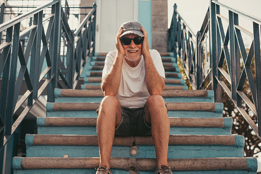 Portrait of senior handsome man with cap and sunglasses looking at camera smiling sitting relaxed in outdoors on a staircase. Carefree elderly man enjoying vacation or retirement