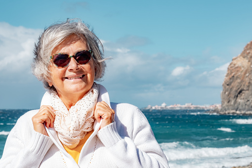Portrait of happy senior white-haired woman in outdoors enjoying sea vacation, elderly smiling lady in a windy day at the beach looking at camera