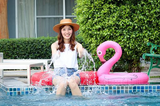Beautiful young Asian woman in summer hat has a great time with outdoor activity while sitting near blue water pool, cheerful female with water splashes has fun, happy girl relaxing by the pool.