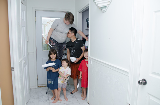Portrait of pregnant woman painting walls while standing of a three-step stool in the corridor of the house.\nFather is kissing the mother's belly and the three kids are facing the camera