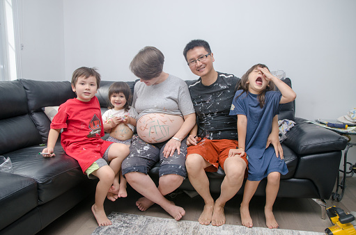 Portrait of happy multiracial family sitting in the black sofa of living room with pregnant mother's belly having been drawn by kids
