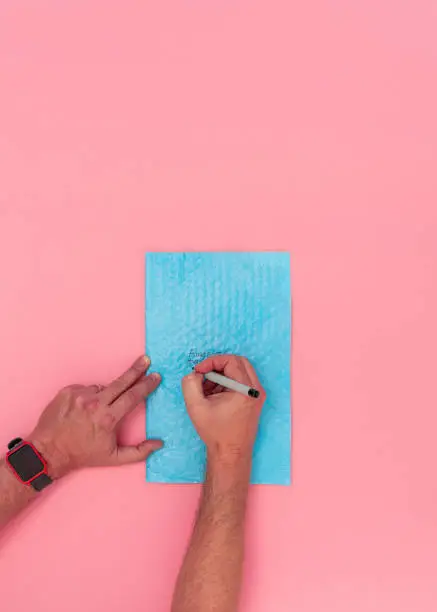 Overhead view of caucasian adult male hands writing a shipping address on a blue bubble packing envelope. Pink background with room for copy space.